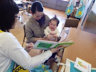母親に抱きかかえられた赤ちゃんに保母さんが絵本を読み聞かせている写真