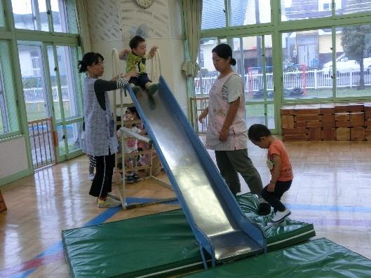 地域交流保育で子供たちが保育士の補助の下園内の滑り台で遊んでいる写真