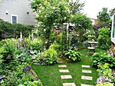 （写真）青々とした芝に石畳が敷かれ、両端には花壇と鉢植えが置かれています。奥には花のアーチがあり、その横にはガーデンテーブルとチェアが置かれ、大変おしゃれなお庭です。