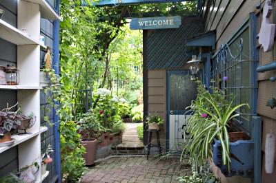 （写真）青い壁には鉢植えや小物が置かれ、上からウエルカムボードとランプがつりさげられています。奥には緑に輝くお庭が見えます。おしゃれなお店の出入り口の様です。