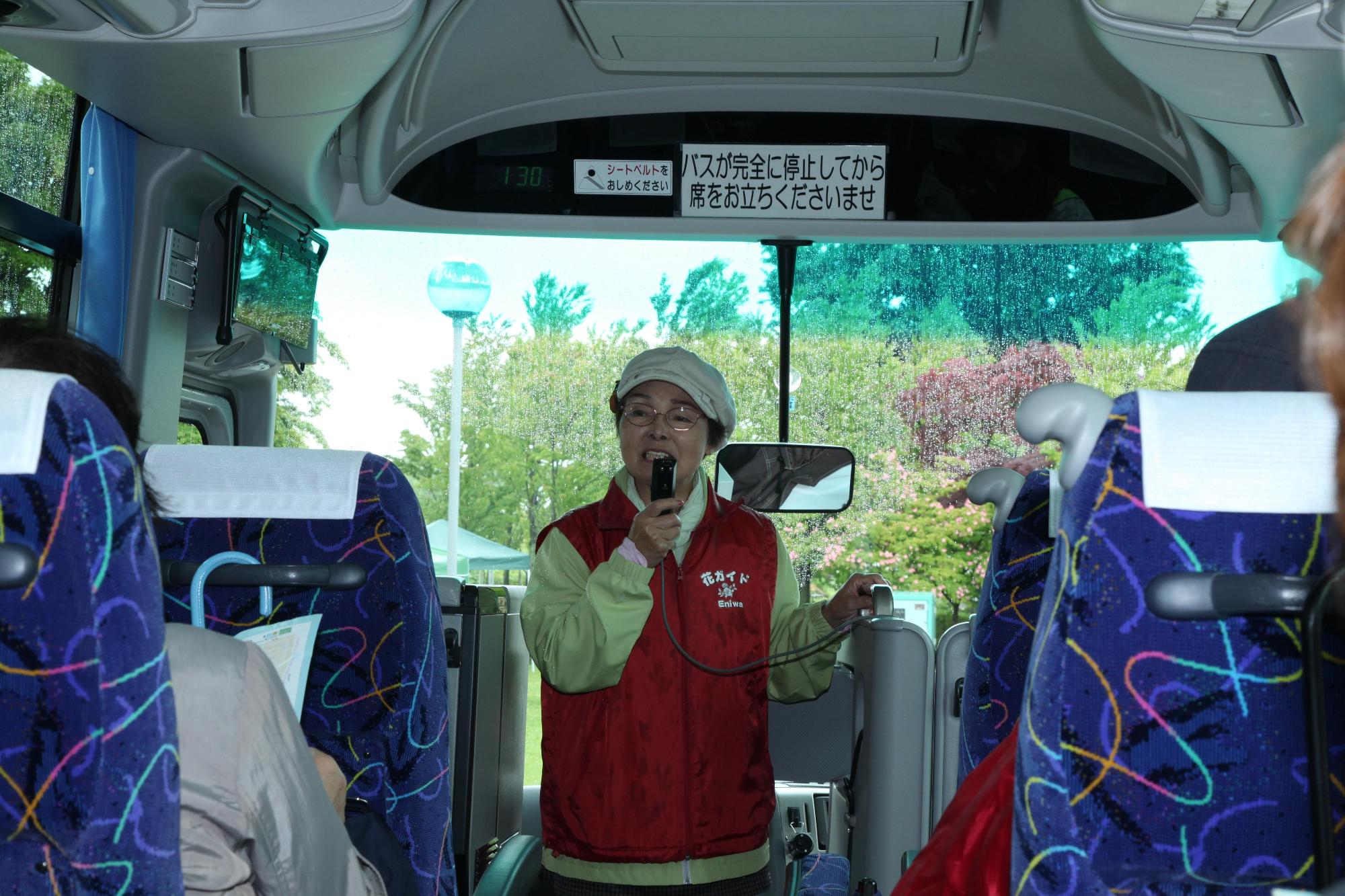 ガーデンツアーのバス内で「花ガイド」とプリントされた赤いベストを着て、ガイドを行う女性の写真
