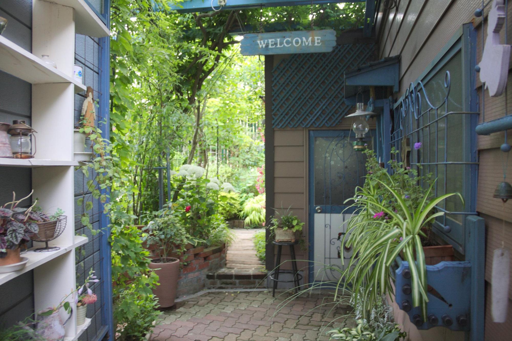 （写真）青い壁のおうちの玄関前にはおしゃれな小物と草木が置かれ、奥には緑に輝くお庭が見えます。