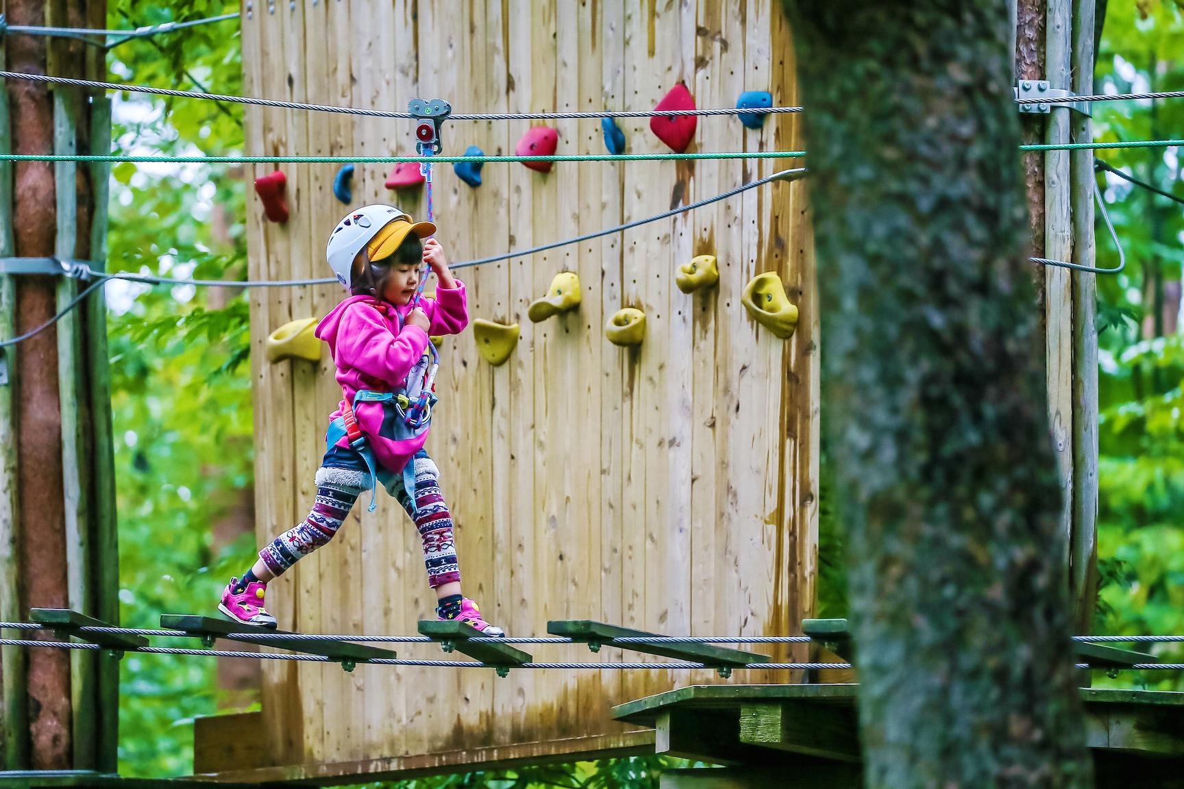 木と木の間に張られたトリムをロープをつかみながら懸命に渡るヘルメットを付けた女の子の写真。奥にはボルダリングの遊具が見えます。