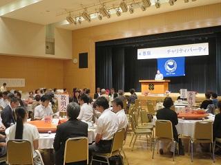 テーブルを囲む出席者とステージ上の原田市長を写した会場の写真