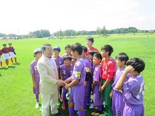 芝生の上で少年サッカーチーム藤枝トレセンのメンバーと握手をしながら声をかける原田市長の写真