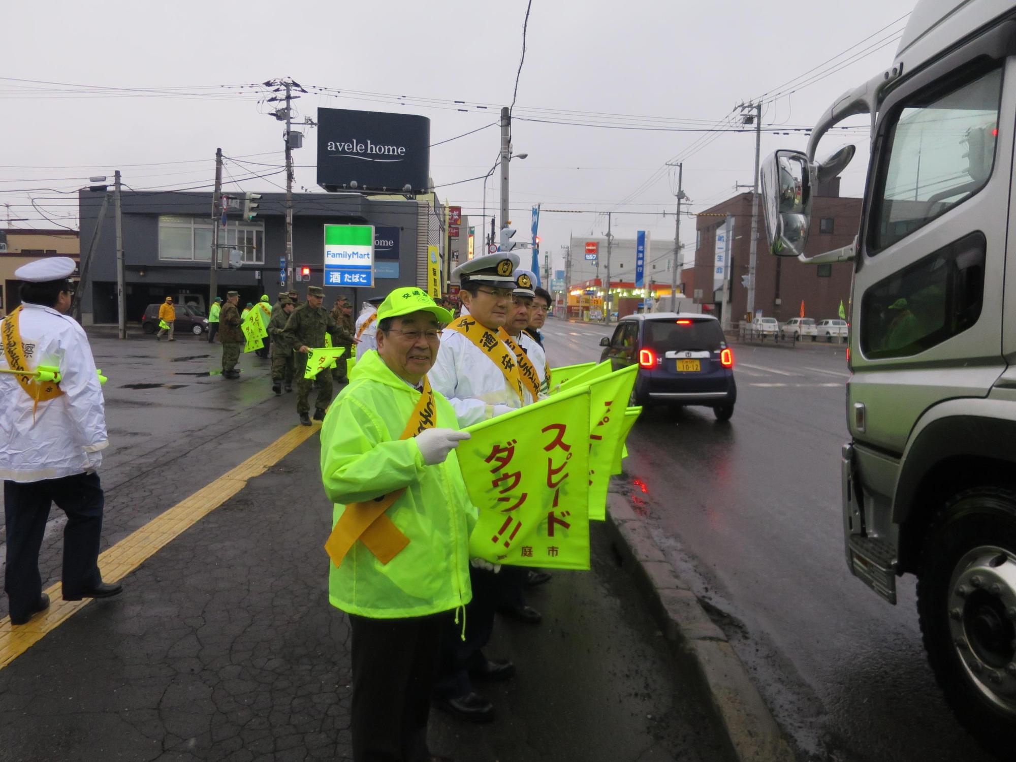 沿道にてスピードダウンの旗を手に通行する車に呼び掛けて原田市長の写真