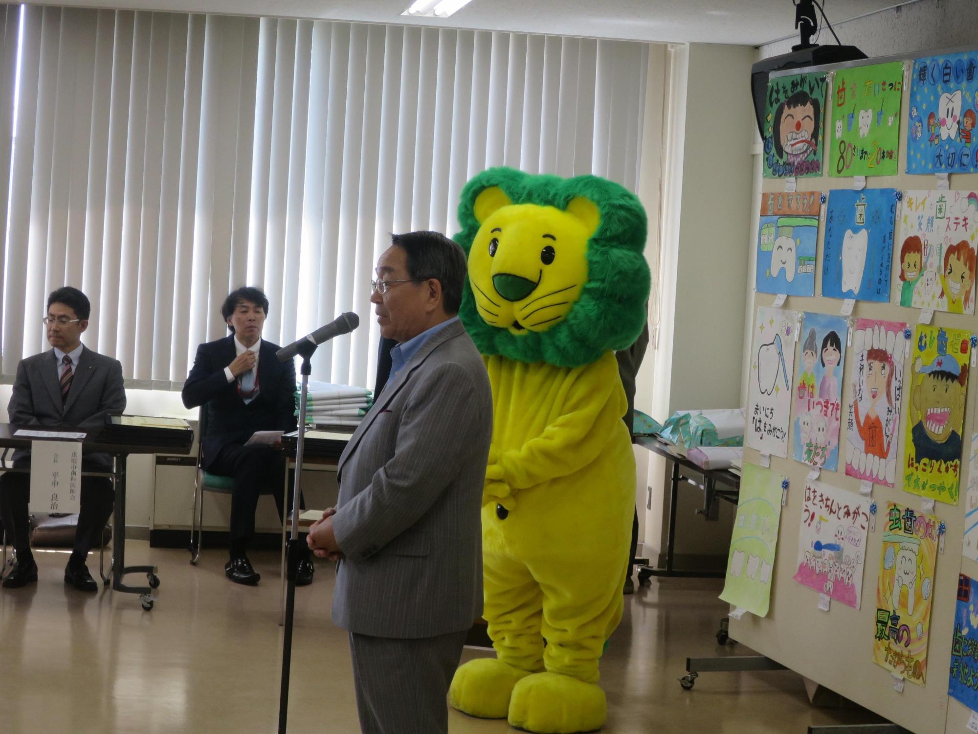 会場で挨拶をする原田市長とマスコットキャラクターのライオンくんの写真