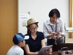 親子がiパッドを見ながら学んでいる隣で、スマートフォンを持ちながら先生が教えている写真。